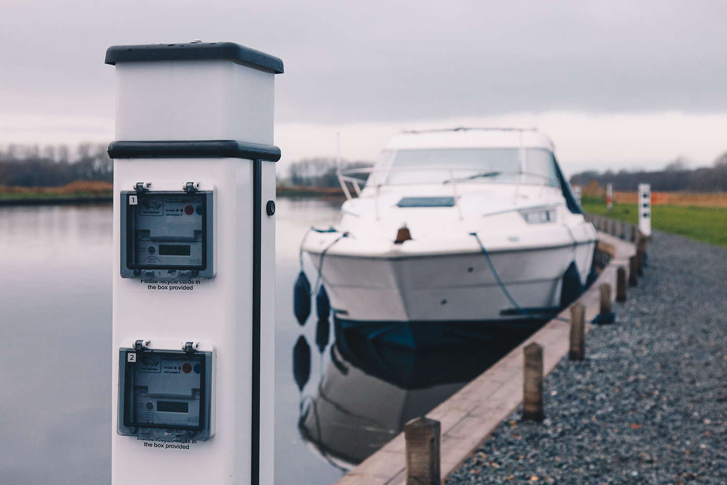 A close-up image of a metal electric charging point on the Broads, with a motor cruiser visible in the distance