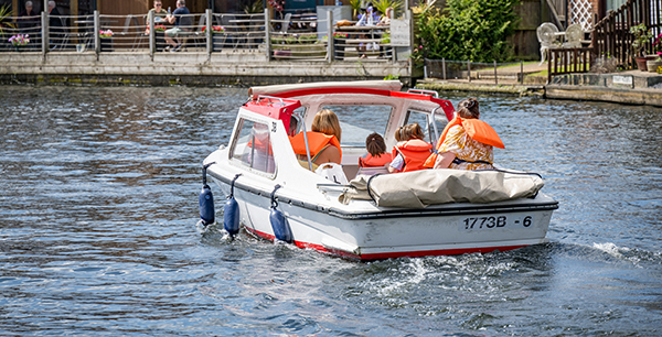 A small white and red day boat carrying four people, all wearing bright orange life jackets, cruises along a river. Two adults and two children sit at the back, enjoying the ride. In the background, a riverside café with people sitting at tables and a wooden deck is visible