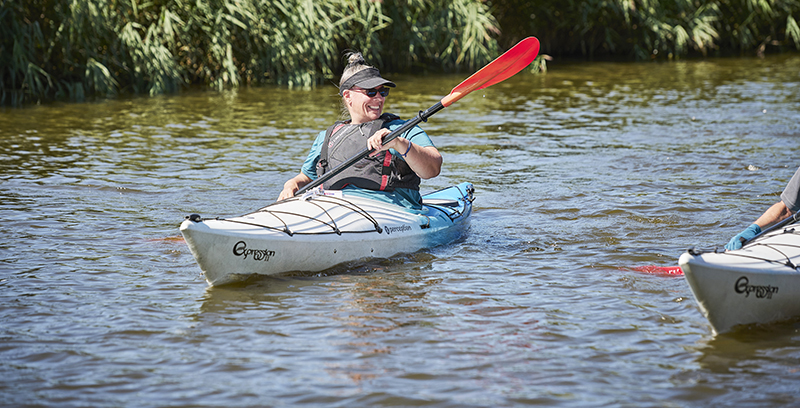 An image showing a person kayaking on a body of water, wearing a life jacket and sunglasses while holding a red paddle. They appear to be enjoying the activity, smiling as they interact with another kayaker. The kayak is branded "Pyranha" and the setting includes lush green vegetation in the background