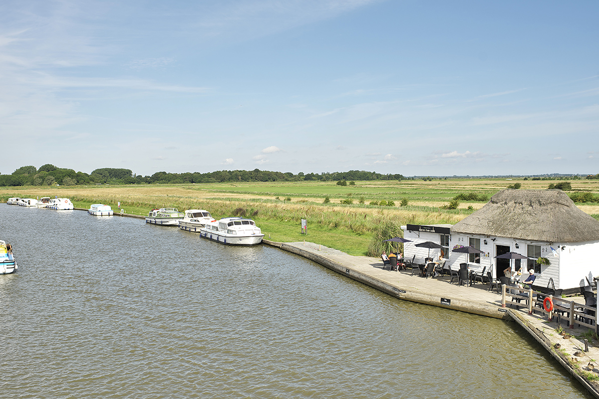 This image shows a serene riverside scene at Acle Bridge, featuring a calm waterway lined with small white boats moored along the bank. To the right, there's a thatched-roof building with outdoor seating, possibly a café or pub, where people are sitting under umbrellas. The surrounding landscape includes open fields with tall grass and trees in the distance, under a bright blue sky with scattered clouds.