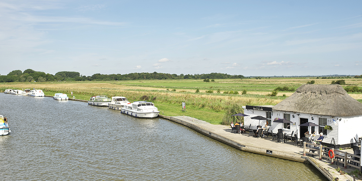 This image shows a serene riverside scene at Acle Bridge, featuring a calm waterway lined with small white boats moored along the bank. To the right, there's a thatched-roof building with outdoor seating, possibly a café or pub, where people are sitting under umbrellas. The surrounding landscape includes open fields with tall grass and trees in the distance, under a bright blue sky with scattered clouds.