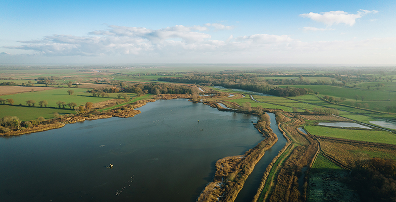 An image shows an aerial view of the Broads’ wetland landscape. A large body of water is surrounded by patches of green fields, marshes, and winding water channels. The horizon stretches far, under a partly cloudy sky, giving the scene a peaceful and expansive feel.