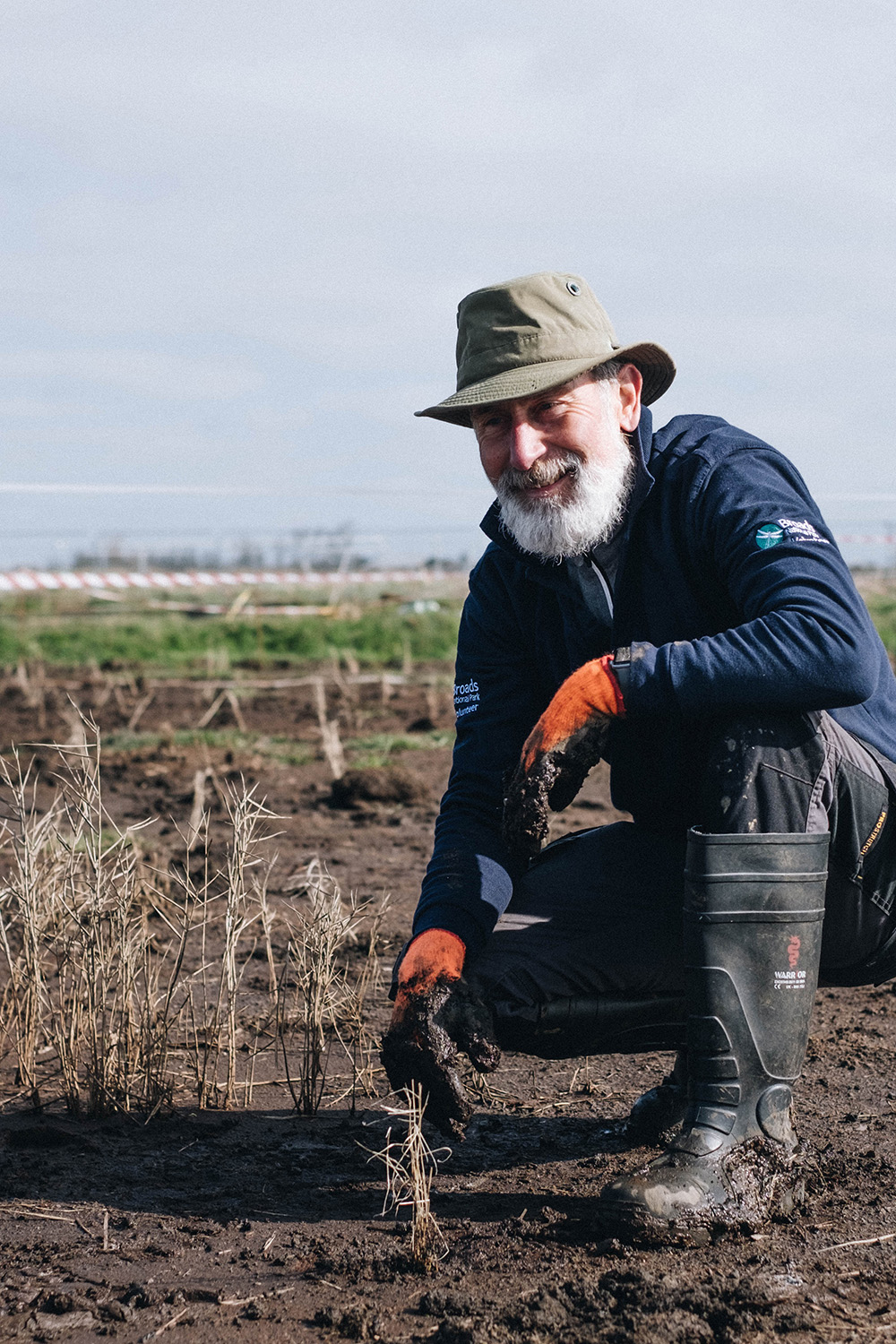 A male volunteer crouching in a field wearing a Broads Authority uniform and a pair of rubber gloves, he appears to be preparing the ground to plant up a plug plant