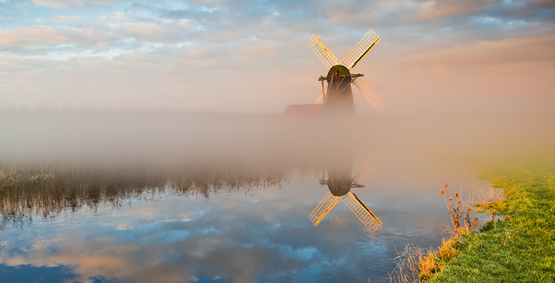 An image showing a black mill surrounded by mist infront of a body of water, the mist and sky are toned with attractive pastel blue and pink colours.