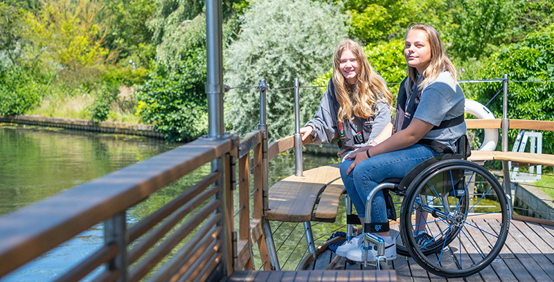 Two young women sitting on a wooden platform or dock near a body of water surrounded by greenery. One of the women is in a wheelchair, and they both appear to be enjoying the outdoors. The setting looks peaceful, with trees and calm water in the background.