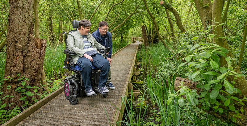 Two people enjoy the accessible boardwalk at Barton Broad, one is a woman in a wheelchair accompanied by a man walking alongside her. They are surrounded by lush green vegetation and appear to have spotted something interesting to look at in the nearby water.