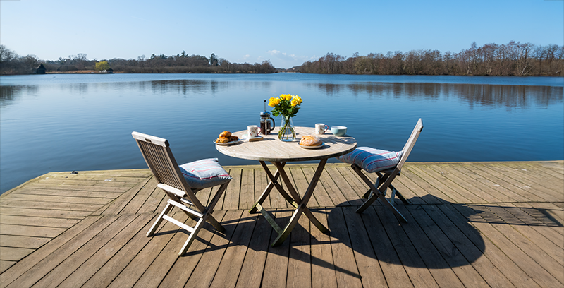 A wooden deck overlooks one of the Broads, with a table set for breakfast with flowers, coffee, pastries, and bread. Two cushioned chairs face the serene water under a clear blue sky.