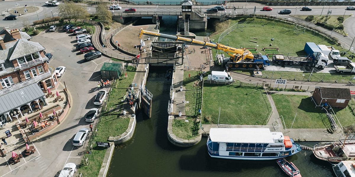 Aerial view of Mutford Lock showing ongoing repair works. Construction equipment and materials are visible near the lock, with workers actively involved in the maintenance. The lock connects Oulton Broad to Lake Lothing, surrounded by moored boats and a marina in the background. Nearby buildings, a parking area, and a busy road with cars are also visible.