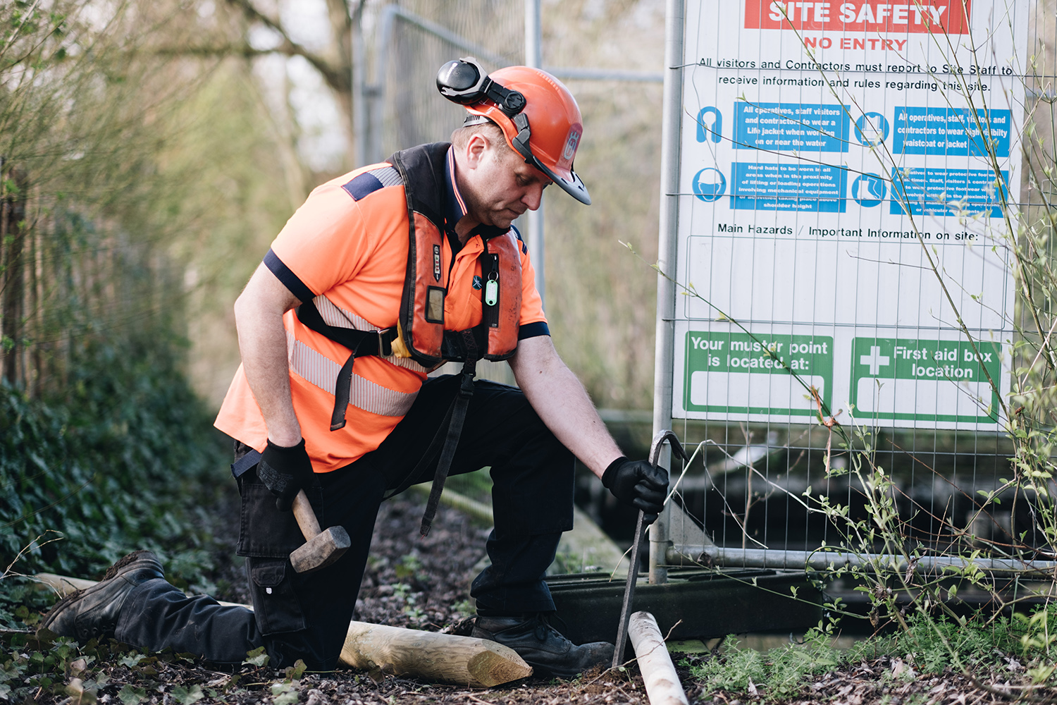 A Broads Authority worker wearing orange PPE uses a hammer to hit something into the ground next to some metal HERAS fencing