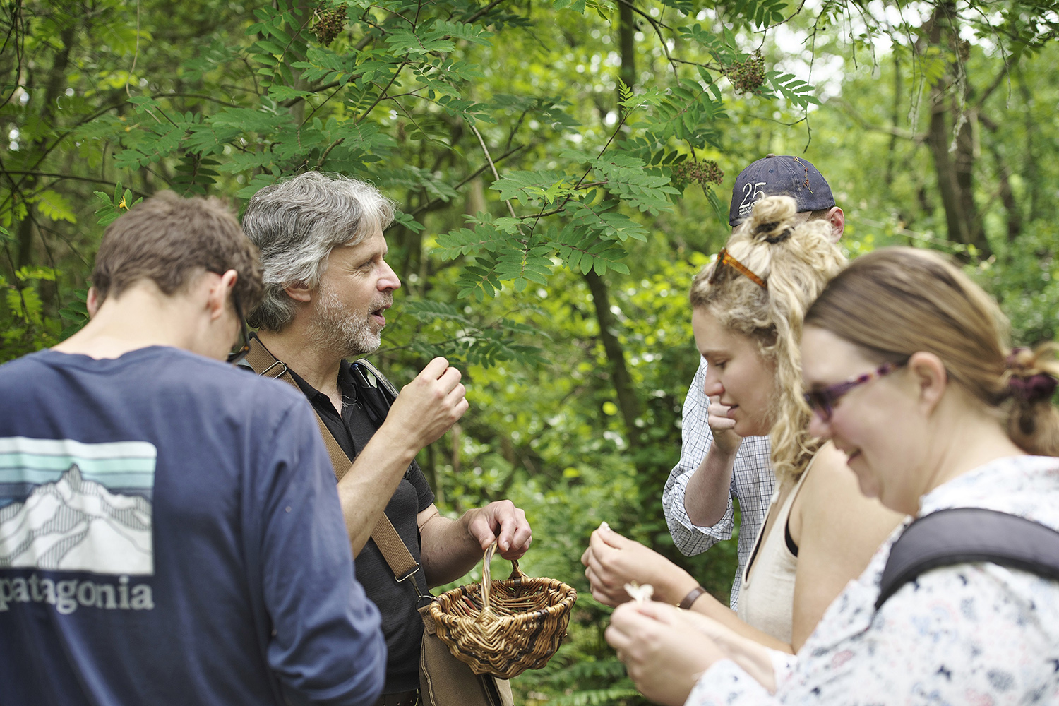 A group of people is gathered in a lush, green forest during a foraging event. A bearded man with gray hair, wearing a black shirt and a leather strap across his chest, appears to be leading the activity, holding a small basket and speaking to the group. The participants, dressed casually, are closely examining and tasting foraged plants, appearing engaged and curious. Sunlight filters through the trees, creating a warm and inviting atmosphere.